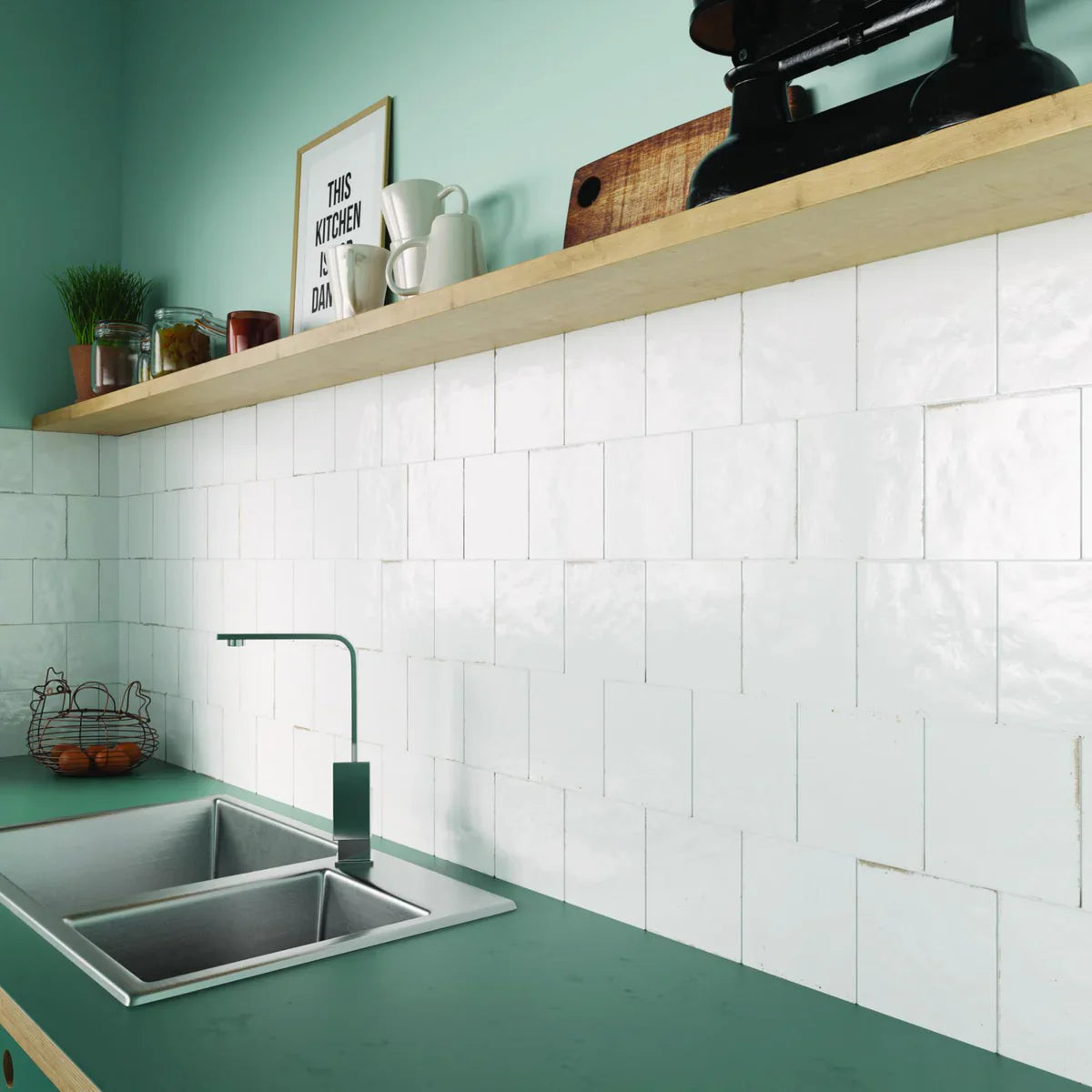 Agadir Niebla 147x147mm white glazed square tiles installed as a kitchen splashback with green countertop, stainless steel sink, and wooden open shelf.