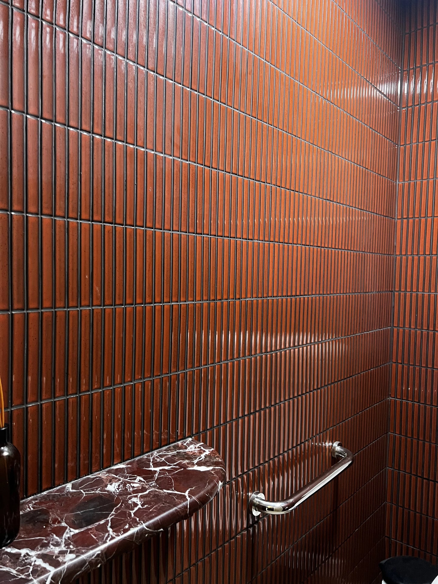 Antique Terracotta Red Speckled Gloss Concave Porcelain Glazed Finger 20x145mm tiles covering a bathroom wall with dark grout, paired with a deep red marble vanity shelf and chrome towel rail.