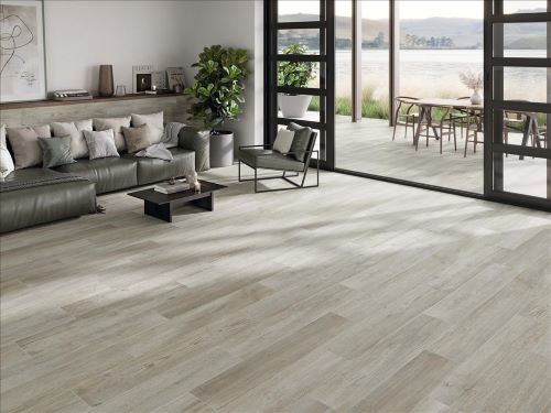 Inout Olson Sky 230x1200mm wood-look grey tiles installed as a living room floor with leather sofa and open glass doors to outdoor dining area.