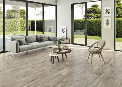 Inout Rigel Grey 150x900mm wood-look porcelain tiles installed as a living room floor, complementing a light grey sofa and rattan chair with garden views beyond.