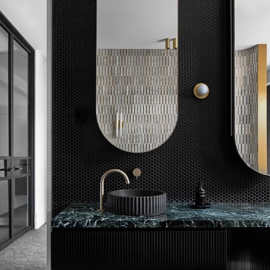 Marlowe Black Matt Mosaic Hexagon 23x23mm tiles covering a dramatic bathroom vanity wall, paired with green marble countertop, arched mirror, and brushed brass faucet.
