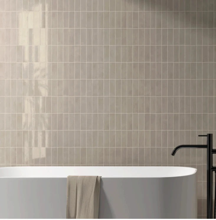 Miyako Morchella Gloss 50x150mm tiles in warm greige covering a full bathroom wall behind a white freestanding bathtub with matte black floor-mounted taps.
