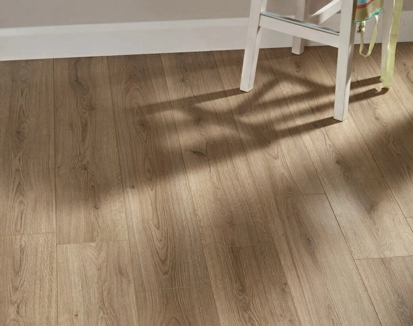 Natural Oak Urban Laminate 12mm Flooring 12mm installed in a bright room with warm sunlight casting shadows from a white chair across the wide-plank wood-effect surface.