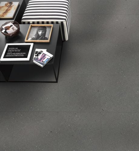 Shellstone Dark Grey Rectified 600x600mm tiles installed as a living room floor beneath a striped ottoman and black coffee table with decorative books and framed portrait.