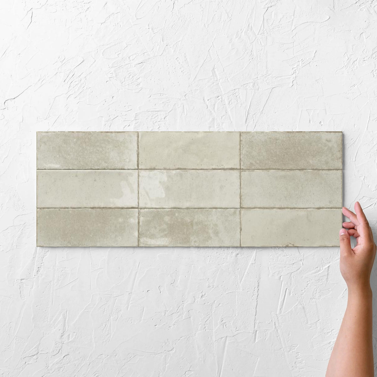 Hands holding an Agadir Ivory 70x280mm tile against a white rendered wall, showing the ivory ultra-gloss surface