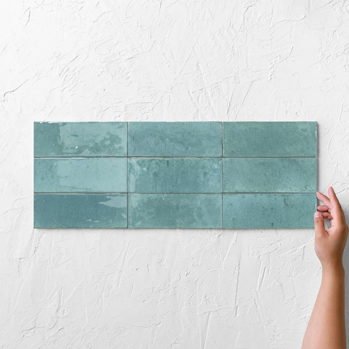 Hands holding an Agadir Turquesa 70x280mm tile against a white rendered wall, showing the turquoise ultra-gloss surface