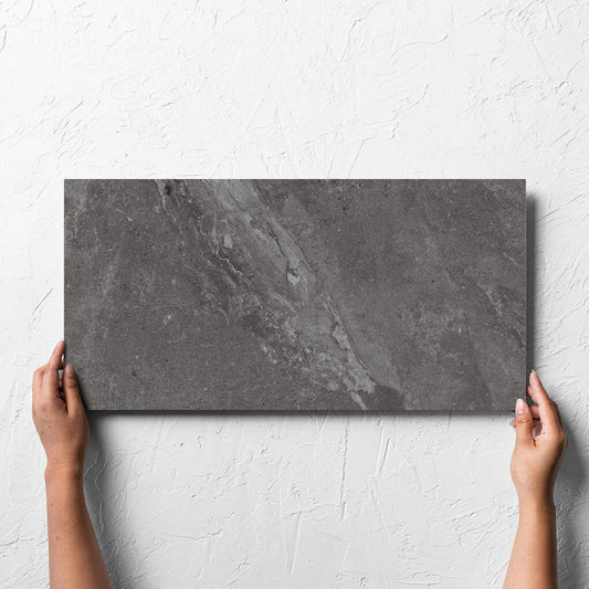Hand holding a single Alpen Stone Smoke 300x600mm tile against a white rendered wall, showing the smoke surface