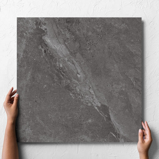 Hand holding a single Alpen Stone Smoke 600x600mm tile against a white rendered wall, showing the smoke surface