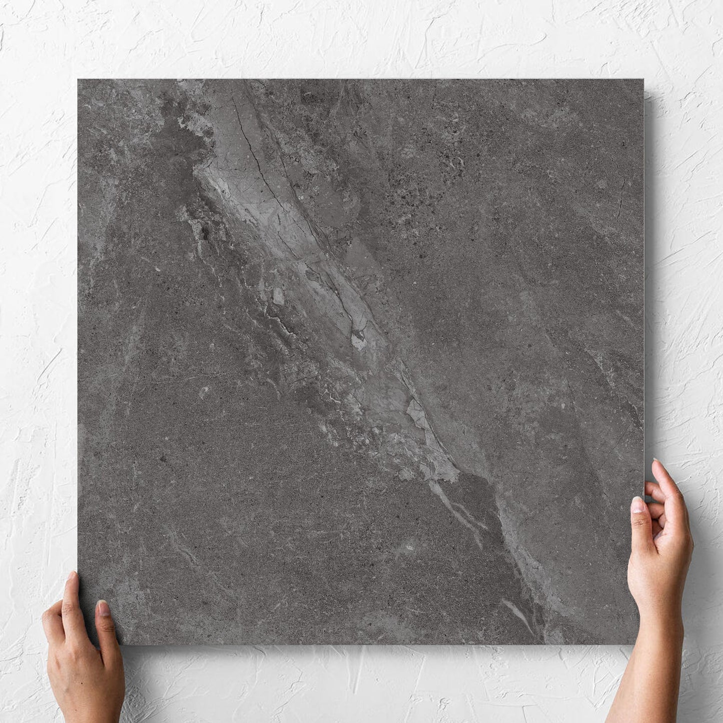 Hand holding a single Alpen Stone Smoke Lappato 600x600mm tile against a white rendered wall, showing the smoke surface