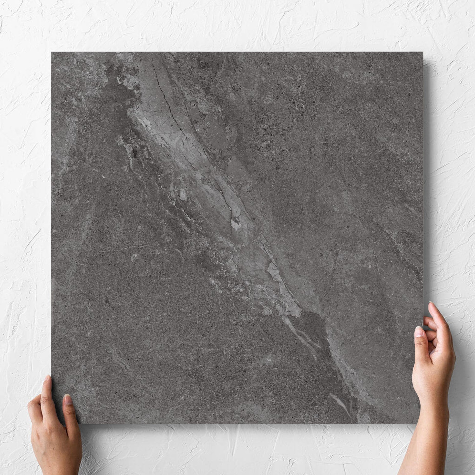 Hand holding a single Alpen Stone Smoke Lappato 600x600mm tile against a white rendered wall, showing the smoke surface