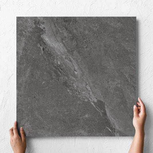 Hand holding a single Alpen Stone Smoke Lappato 600x600mm tile against a white rendered wall, showing the smoke surface