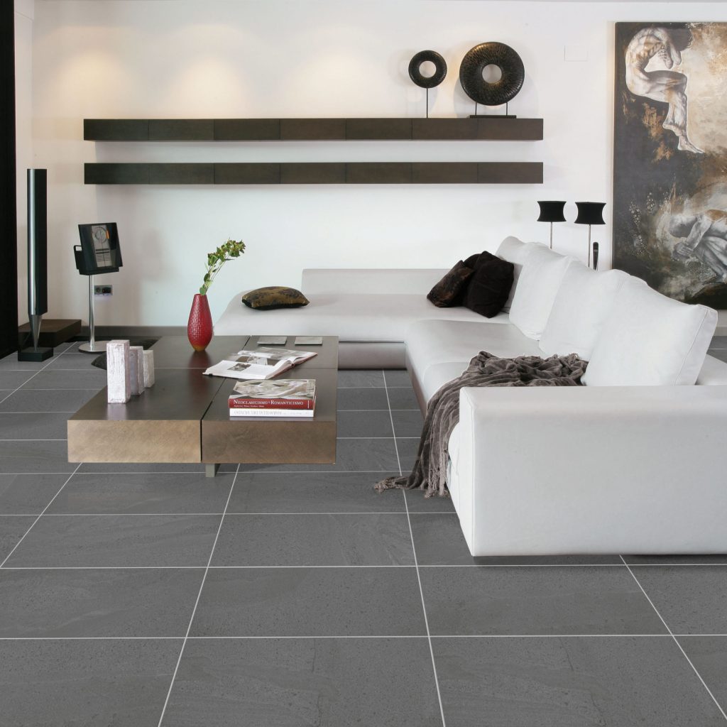 Alps Dark Grey 300x600mm tiles laid as a living room floor, complementing a white sectional sofa, bronze coffee table, and contemporary wall shelving.