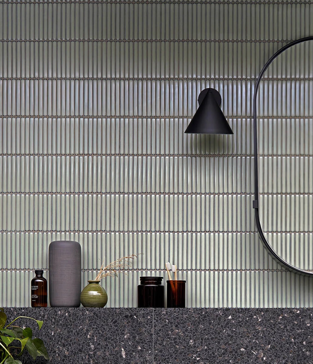 Antique Green Gloss Porcelain Glazed KitKat 12x92mm tiles installed as a full bathroom vanity feature wall with a black wall sconce, round mirror, and dark terrazzo countertop.