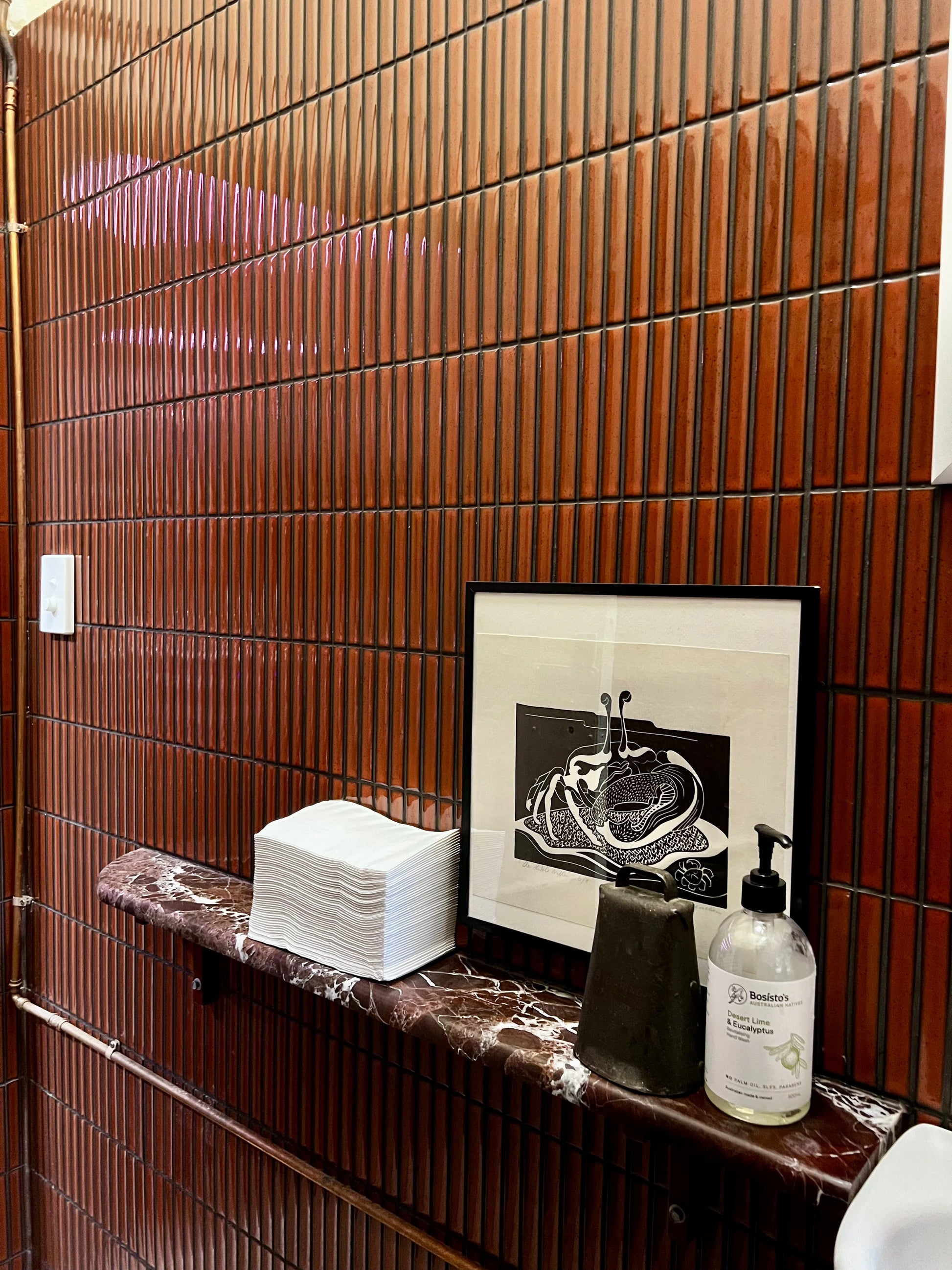 Antique Terracotta Red Speckled Gloss Concave Porcelain Glazed Finger 20x145mm tiles covering a bathroom wall above a dark red marble vanity shelf with hand soap and artwork.