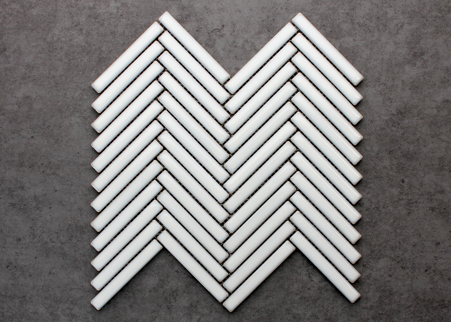 Antique White Gloss Porcelain Glazed Herringbone KitKat 12x92mm mosaic sheet displayed flat, showing glossy white finger tiles arranged in a classic chevron herringbone pattern against a grey concrete background.