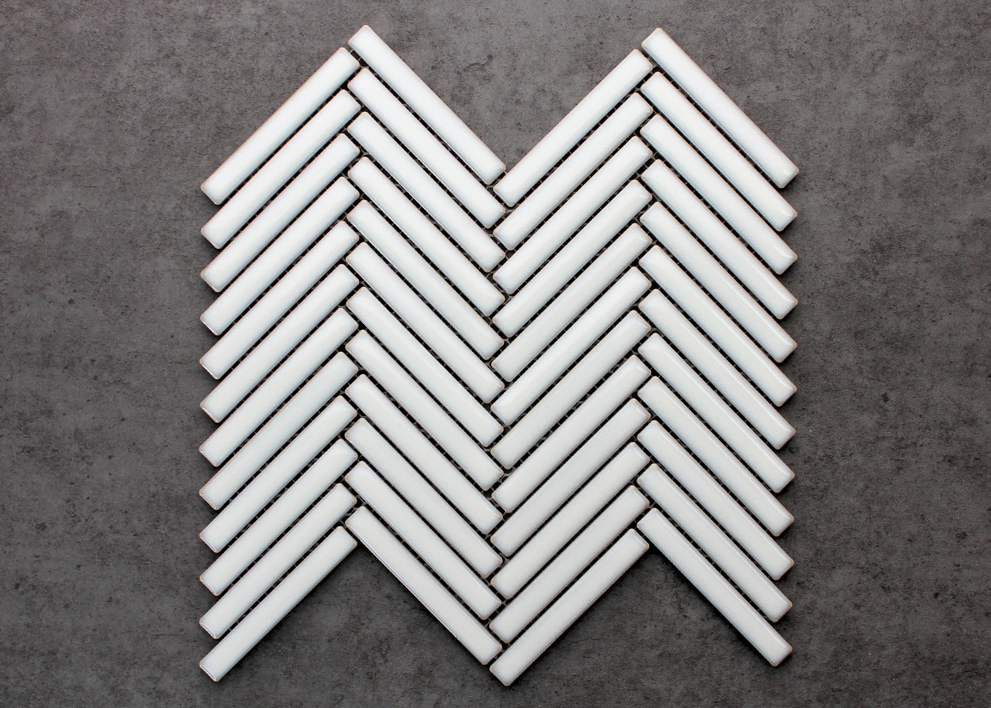 Antique White Gloss Porcelain Glazed Herringbone KitKat 12x92mm mosaic sheet displayed flat, showing glossy white finger tiles arranged in a classic chevron herringbone pattern against a grey concrete background.