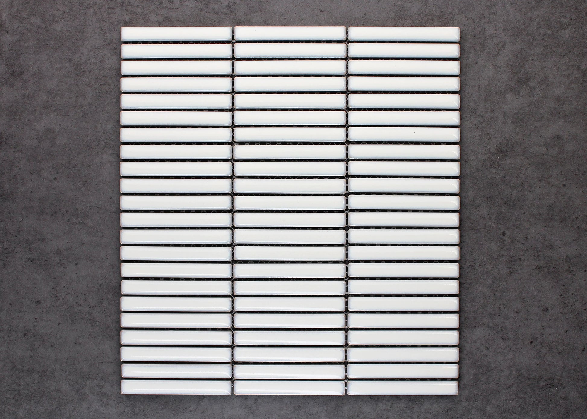 A flat-lay pack shot of Antique White Gloss Porcelain Glazed KitKat 12x92mm tiles arranged in three stacked mesh-mounted columns against a grey concrete background.