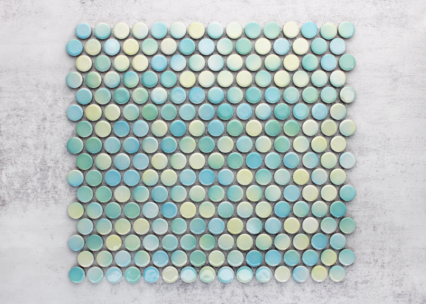 A flat-lay pack shot of Aqua Mix Gloss Diameter Glazed Porcelain Penny Round 19mm tiles arranged in a mesh-backed sheet, displaying a blended mix of aqua, teal, and pale green glossy circular tiles on a grey surface.
