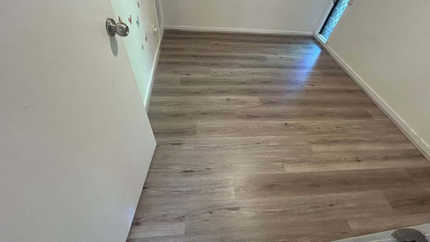 Ash Cliff Elegant SPC Hybrid 6.5mm Flooring 5+1.5mm installed in an empty bedroom with grey-brown wood-look planks, white skirting boards, and natural window light.