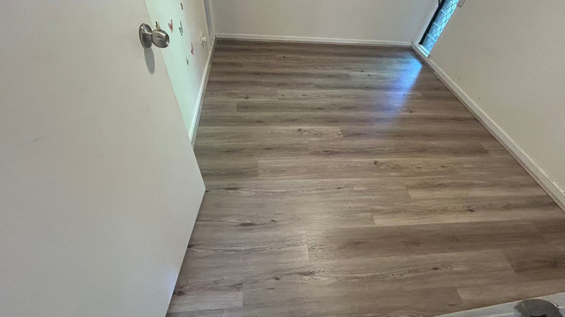 Ash Cliff Elegant SPC Hybrid 6.5mm Flooring 5+1.5mm installed in an empty bedroom with grey-brown wood-look planks, white skirting boards, and natural window light.