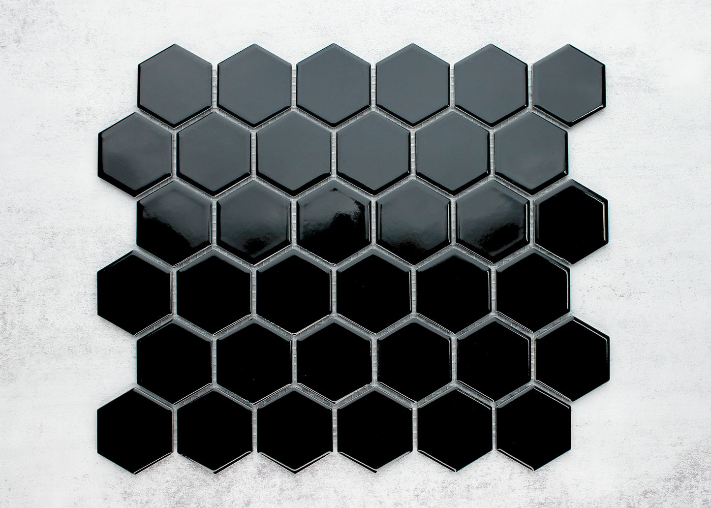 Black Gloss Porcelain Glazed Hexagon 51x59mm tiles arranged in a gradient pattern from dark charcoal to deep gloss black on a light concrete background.