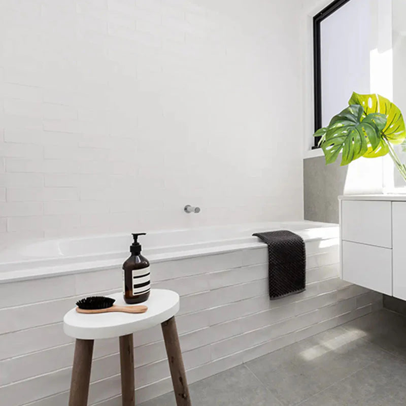 Block White Matt 76x302mm subway tiles lining a minimalist bathroom bathtub surround and feature wall, styled with a timber stool and monstera plant.