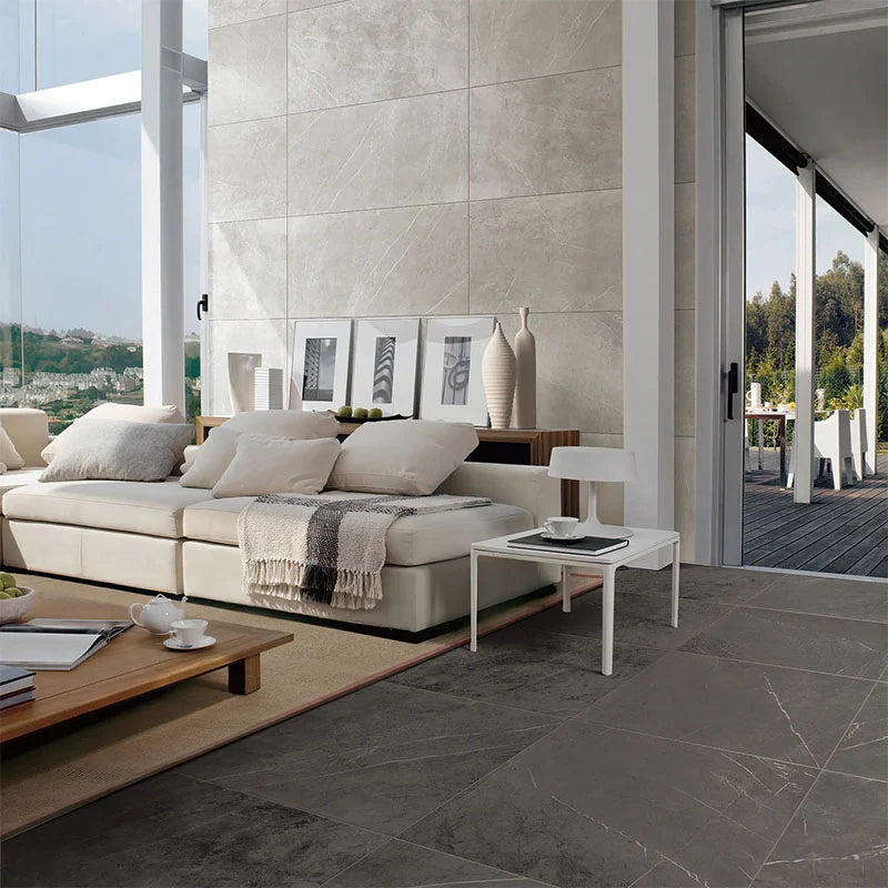 Bracca Dark Grey Lappato Rectified 600x600mm tiles covering a contemporary living room floor, paired with a cream sofa, marble-look feature wall, and open terrace beyond.