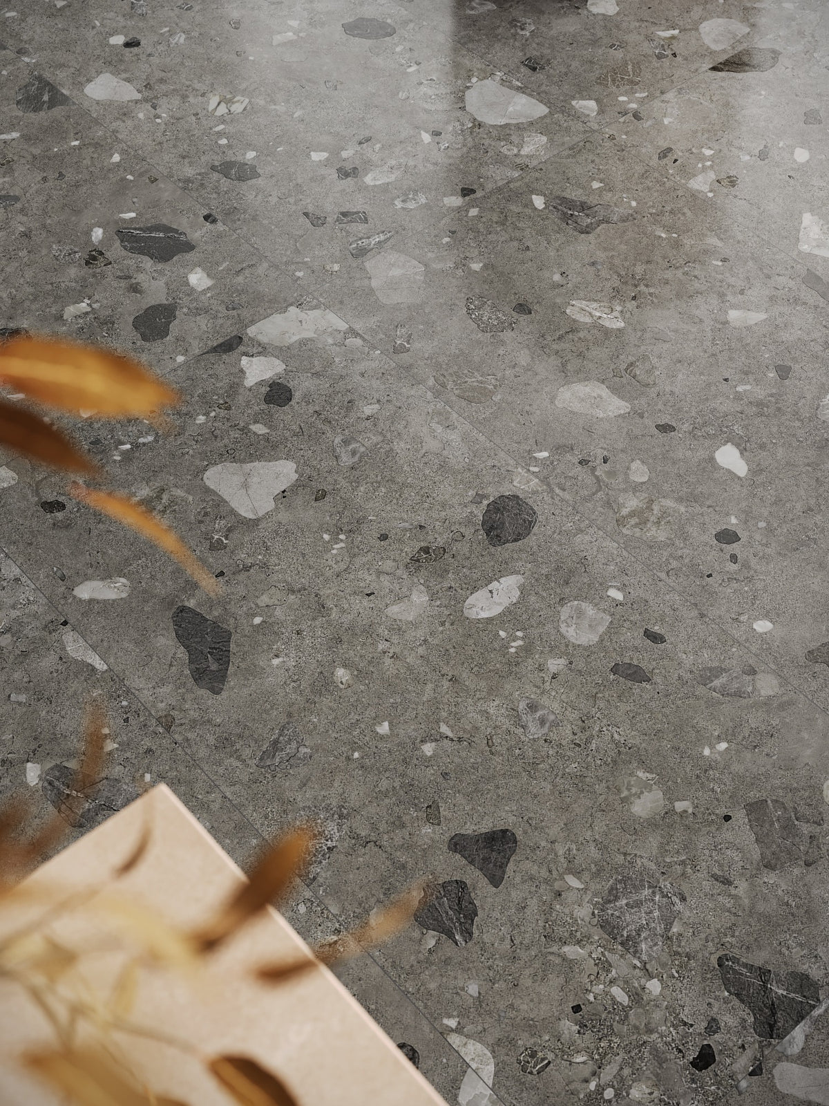 Brescia Iconic Grafite Premium Honed 600x1200mm tile installed as a polished grey terrazzo-style floor with scattered black and white stone fragments, styled with dried grasses and a timber vase.
