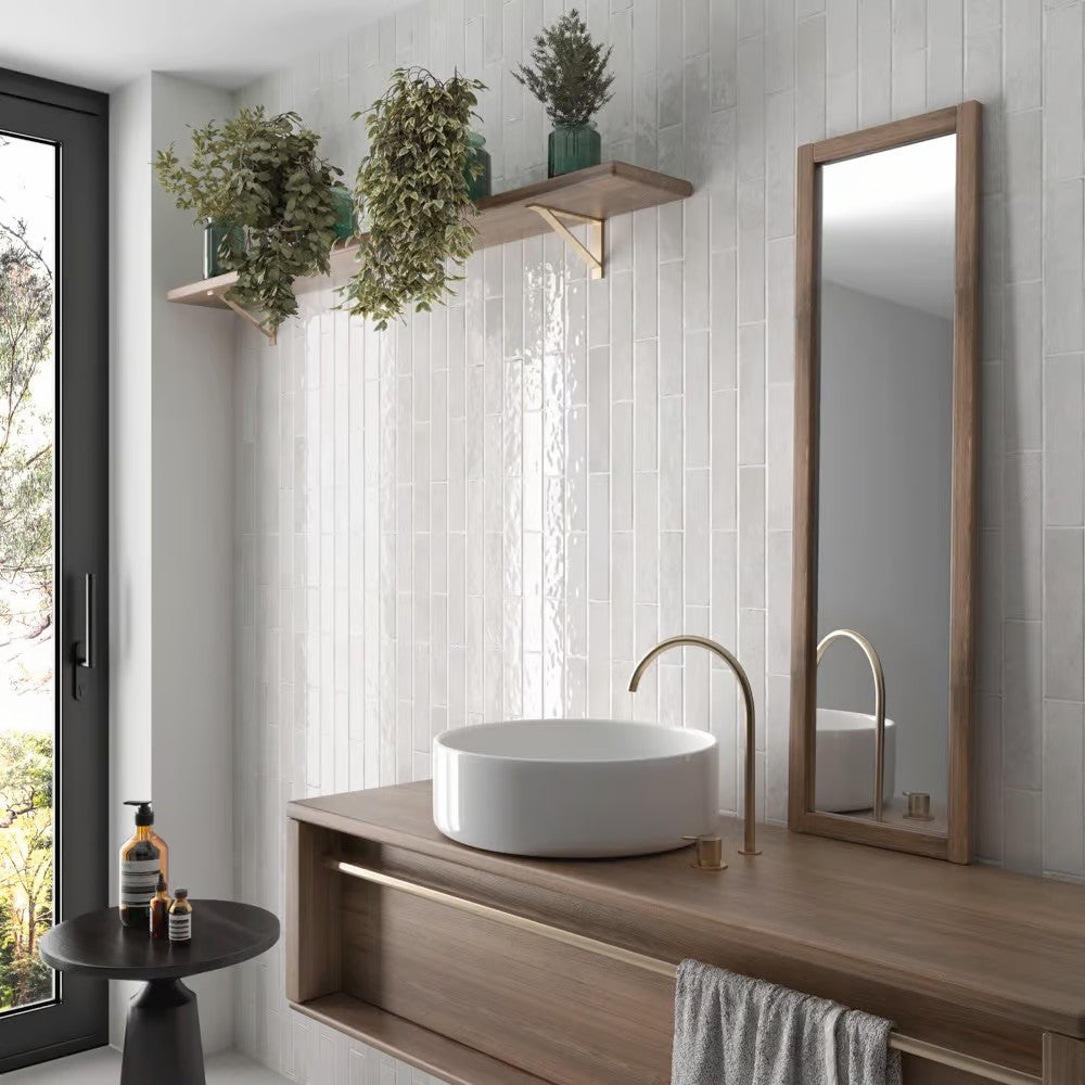 Bricks White Gloss 60x246 tiles installed vertically on a bathroom feature wall behind a timber vanity with vessel sink and wooden floating shelves holding indoor plants.