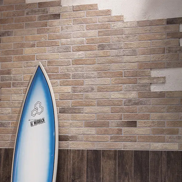 Bristol Rust Brick Tile 60x250mm installed as a feature wall in a coastal-style interior, styled with a blue Al Merrick surfboard leaning against the warm sandy-toned brick-effect tiled wall.
