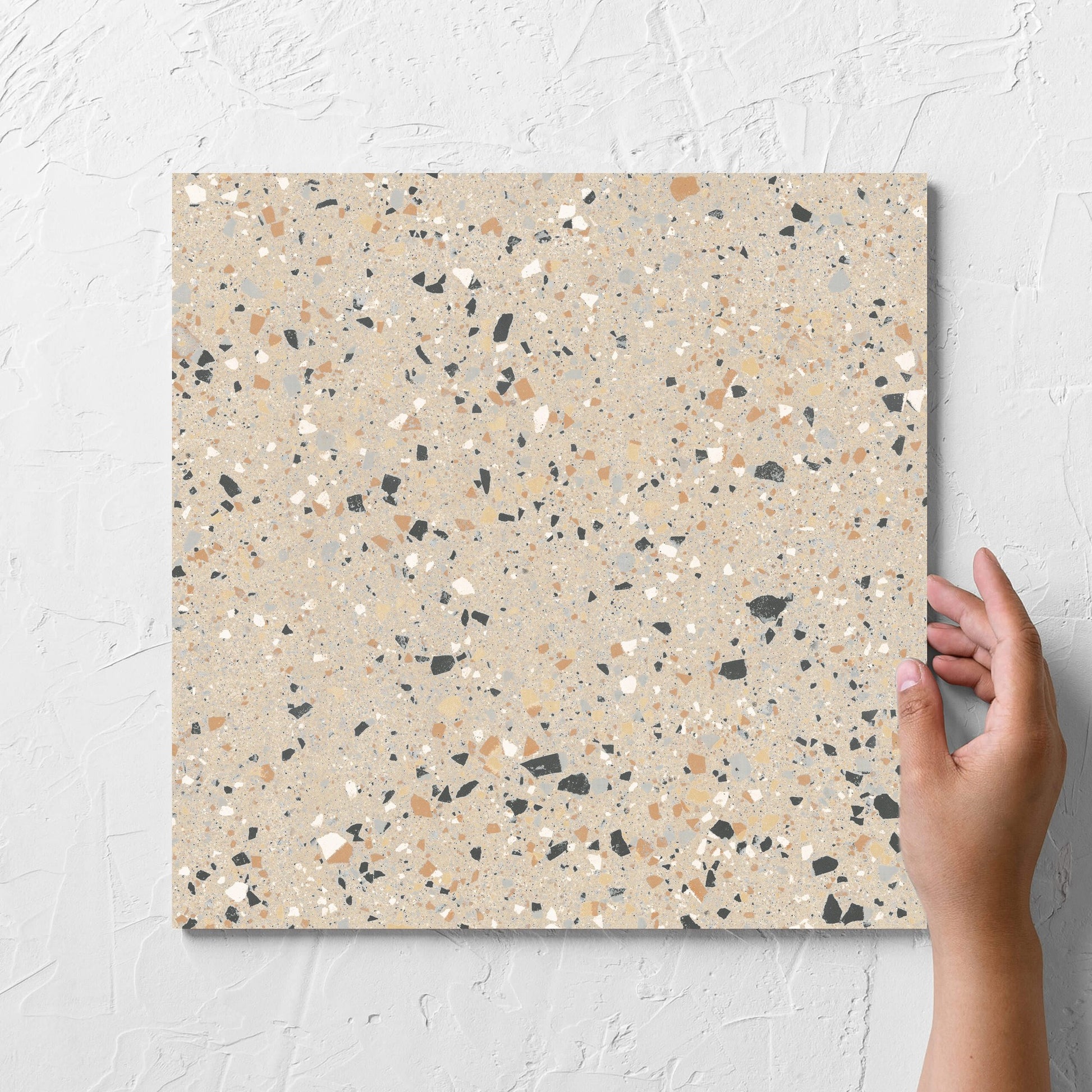 Hand holding a single C Venice 300x300 Matt Beige Terrazzo Look Rectified tile against a white rendered wall, showing the beige terrazzo look surface