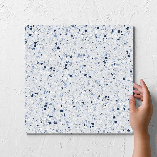 Hand holding a single C Venice 300x300 Matt Blue Terrazzo Look Rectified tile against a white rendered wall, showing the blue terrazzo look surface