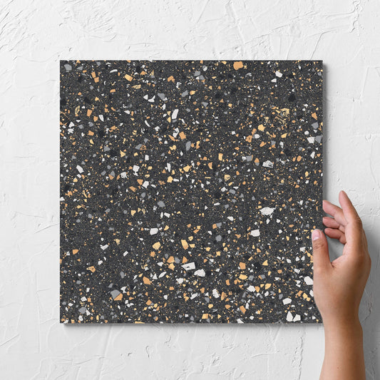 Hand holding a single C Venice 300x300 Matt Graphite Terrazzo Look Rectified tile against a white rendered wall, showing the terrazzo look tiles surface