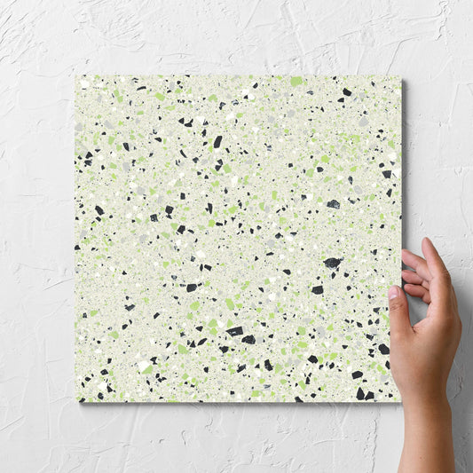 Hand holding a single C Venice 300x300 Matt Green Terrazzo Look Rectified tile against a white rendered wall, showing the tile surface