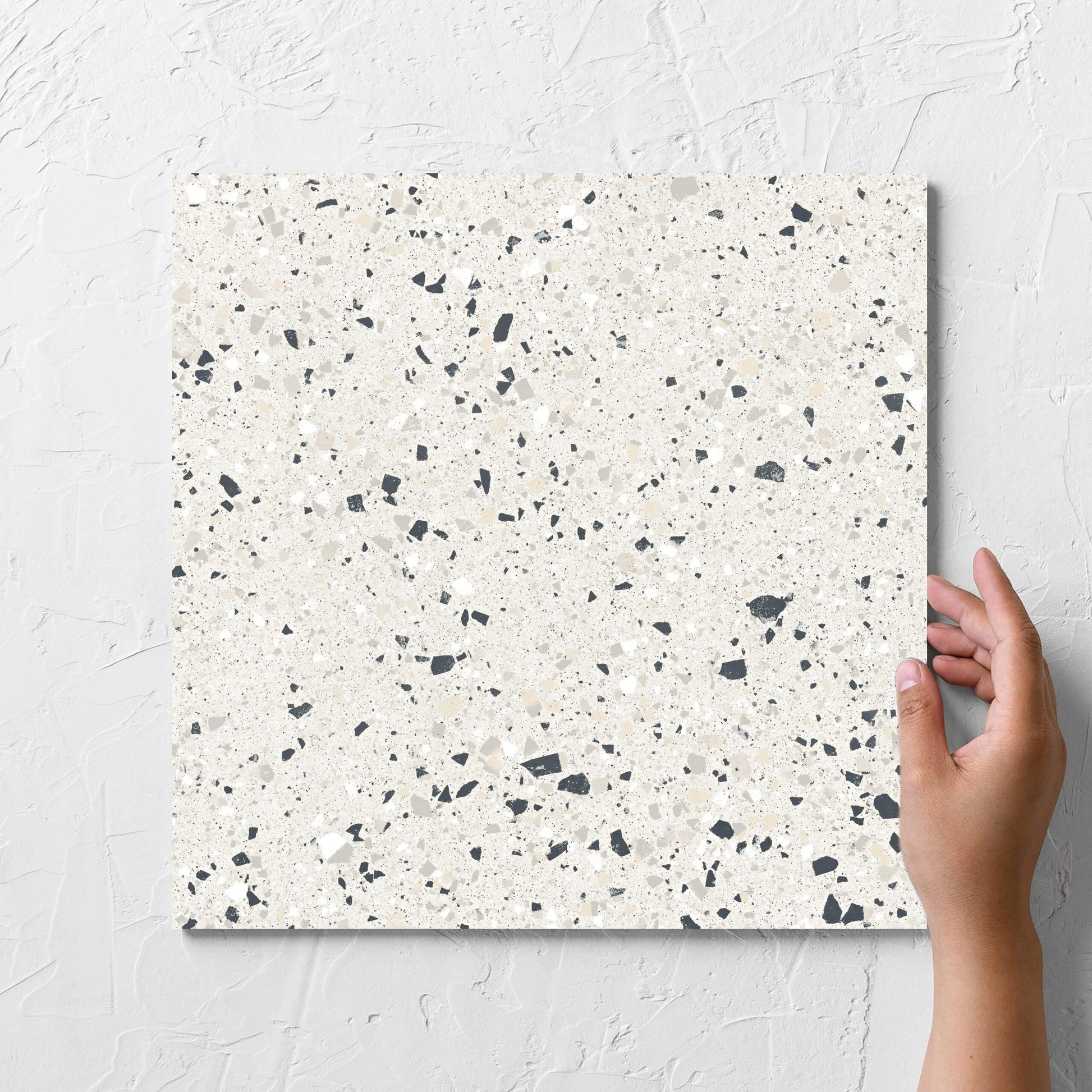 Hand holding a single C Venice 300x300 Matt Silver Terrazzo Look Rectified tile against a white rendered wall, showing the tile surface