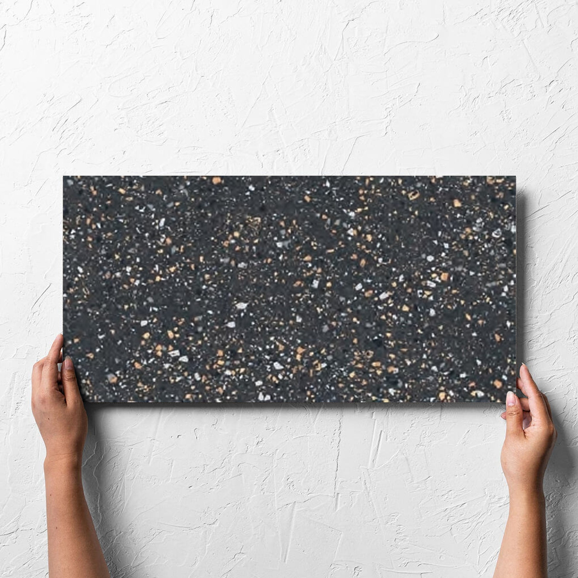 Hand holding a single C Venice 300x600 Matt Graphite Terrazzo Look Rectified tile against a white rendered wall, showing the graphite matt surface