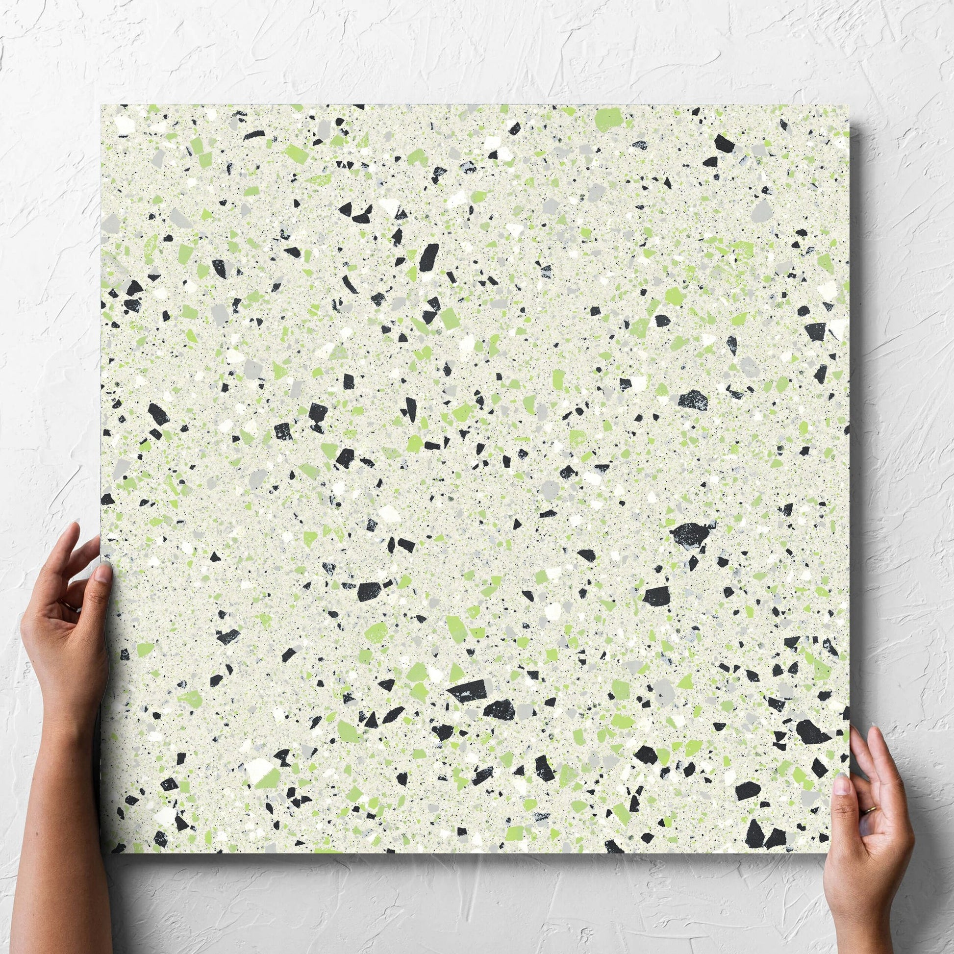 Hand holding a single C Venice 600x600 Matt Green Terrazzo Look Rectified tile against a white rendered wall, showing the tile surface