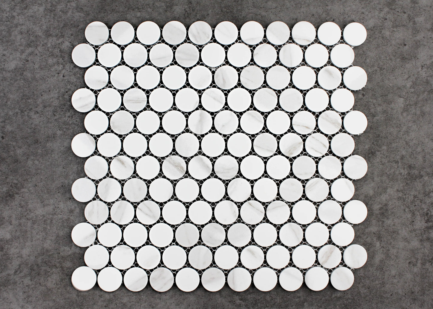 Pack shot of Carrara Look Matte Glazed Porcelain Penny Round 28mm Diameter tiles arranged in a mesh-backed sheet showing white circular discs with subtle grey veining on a dark grey background.
