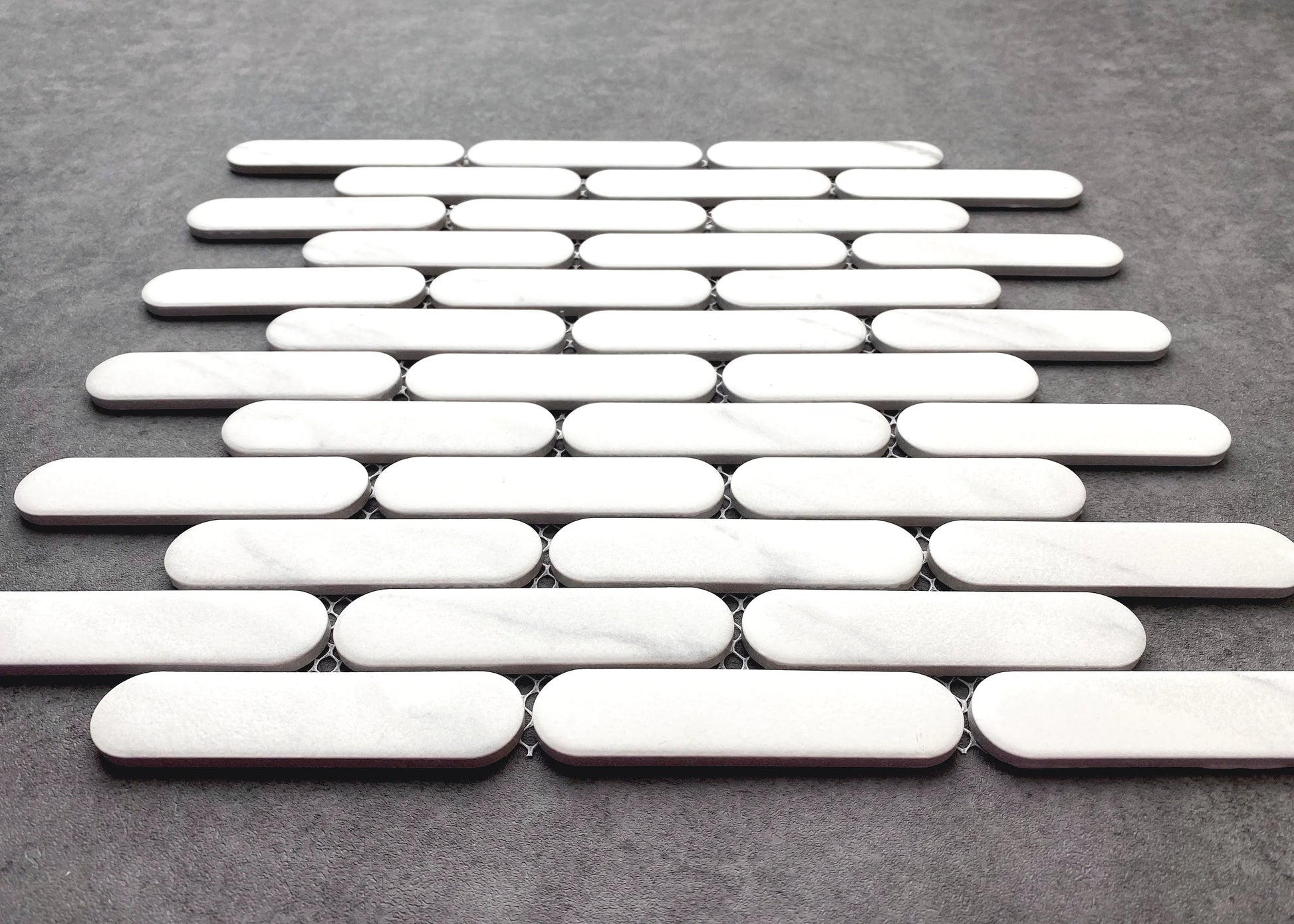 Closeup of Carrara Matte Round Head Finger Glazed Mosaic 23x93mm showing white marble oval tiles arranged in staggered rows on mesh backing against a grey surface.