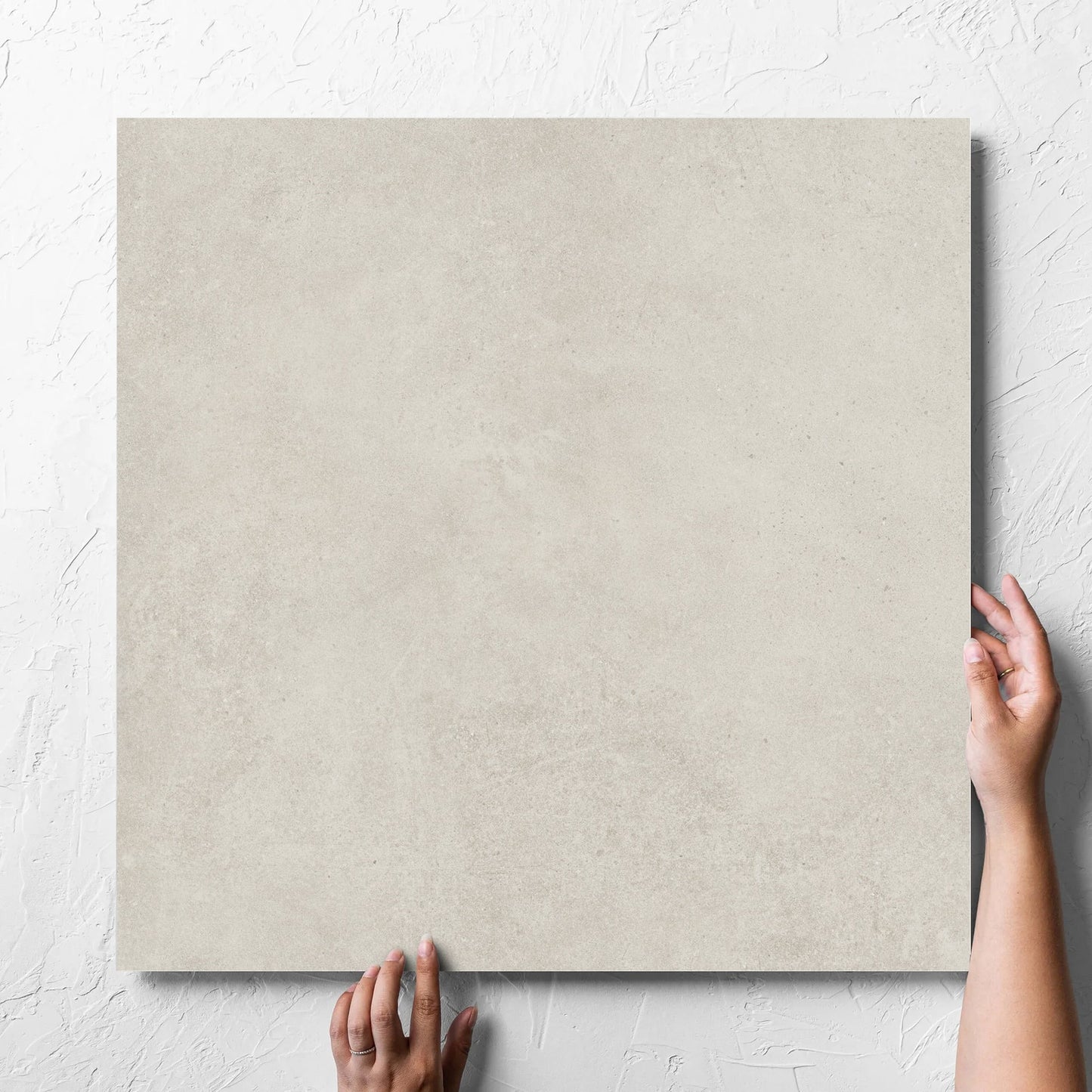 Hand holding a single Cement Light Grey Matt 600x600mm tile against a white rendered wall, showing the matt surface