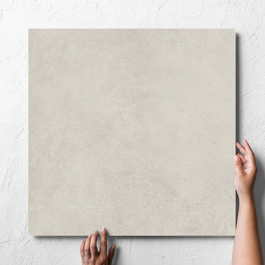 Hand holding a single Cement Light Grey Matt 600x600mm tile against a white rendered wall, showing the matt surface