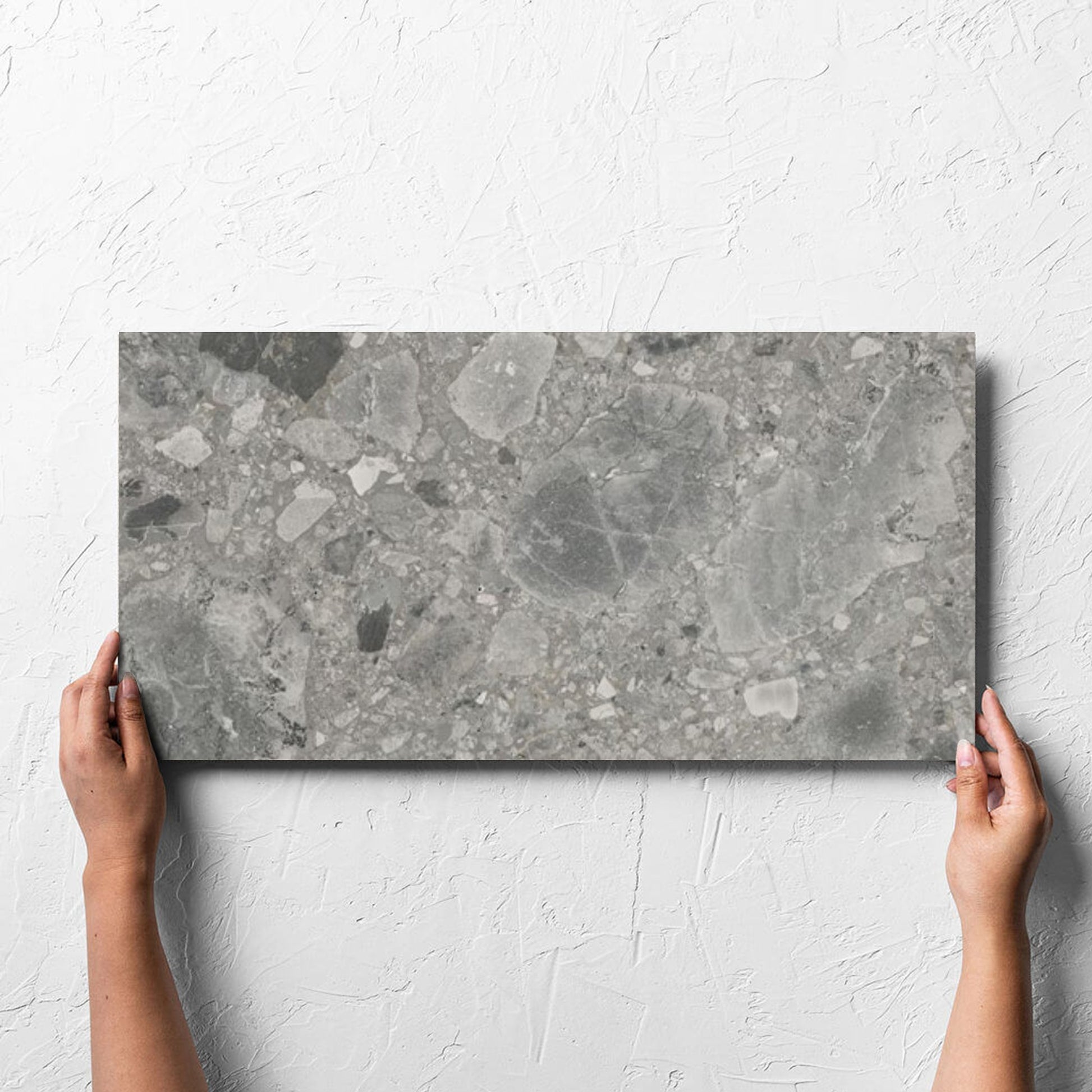 Hand holding a single Ceppo Grey InOut 300x600mm tile against a white rendered wall, showing the tile surface