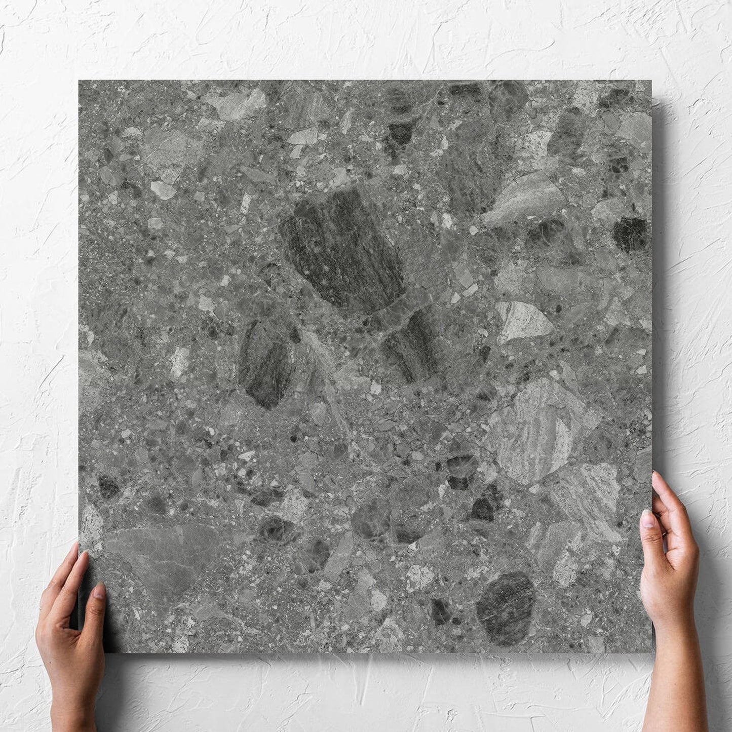 Hand holding a single Ceppo Grey InOut 600x600mm tile against a white rendered wall, showing the tile surface