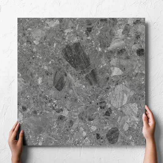 Hand holding a single Ceppo Grey InOut 600x600mm tile against a white rendered wall, showing the tile surface