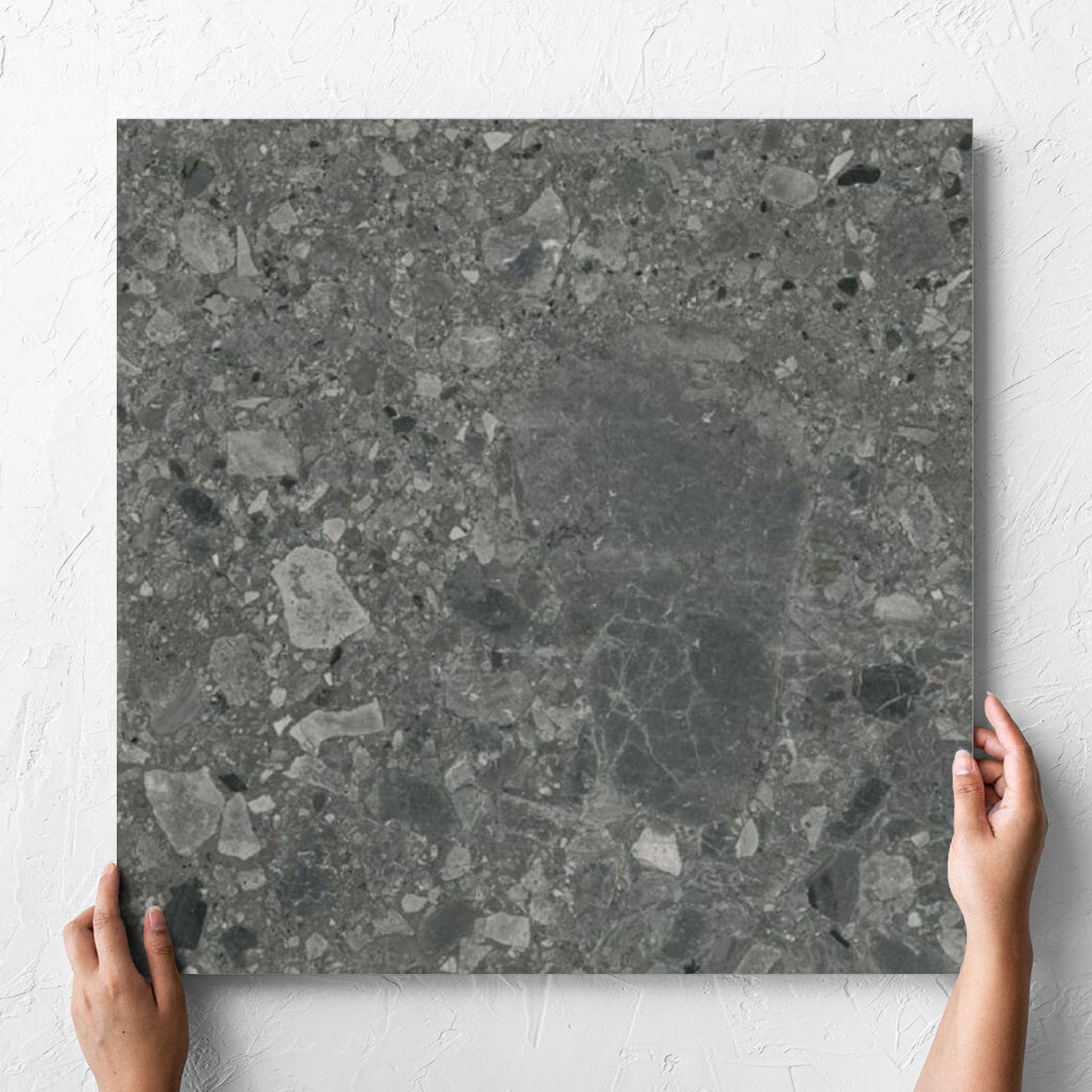 Hand holding a single Ceppo Nero InOut 600x600mm tile against a white rendered wall, showing the tile surface