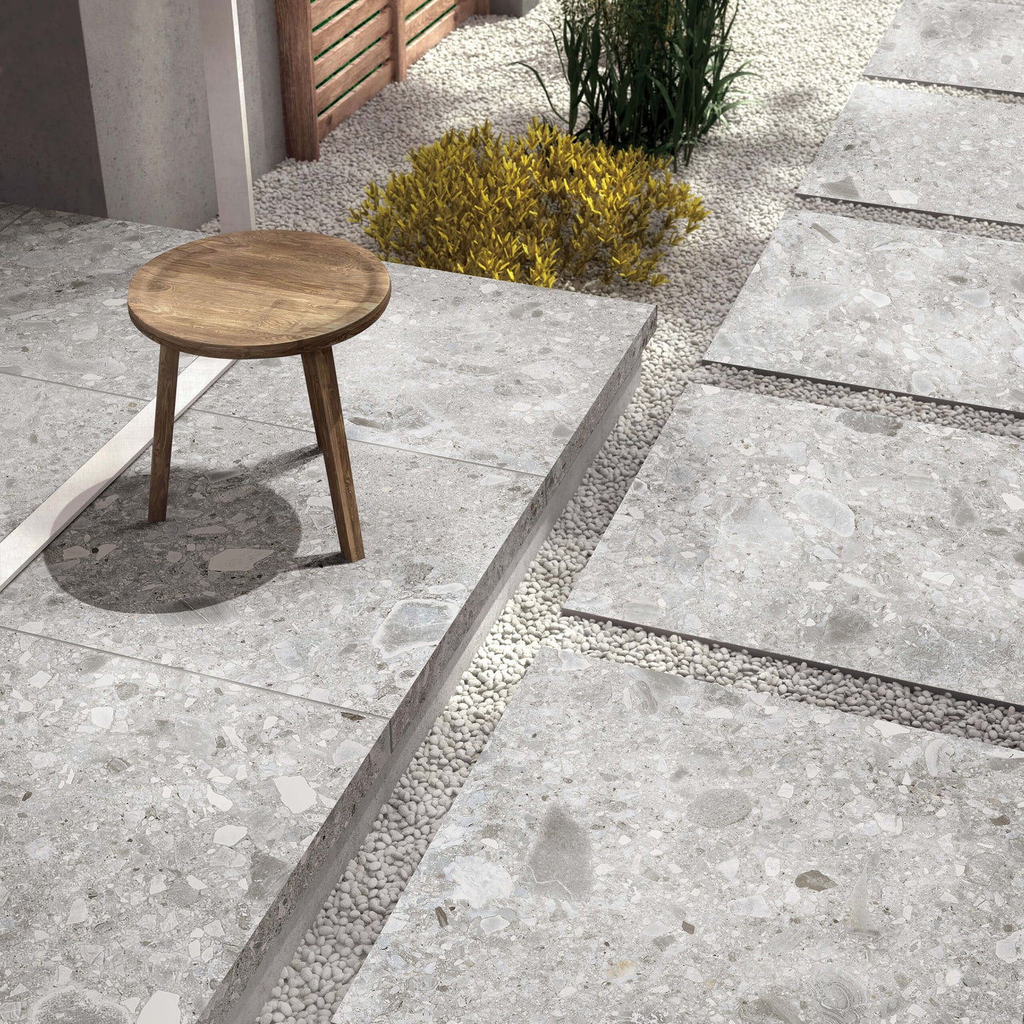 Ceppo Stone InOut 600x600mm grey terrazzo-effect porcelain tiles laid as raised outdoor patio stepping platforms separated by white pebble gravel channels, styled with a wooden stool and ornamental shrubs.