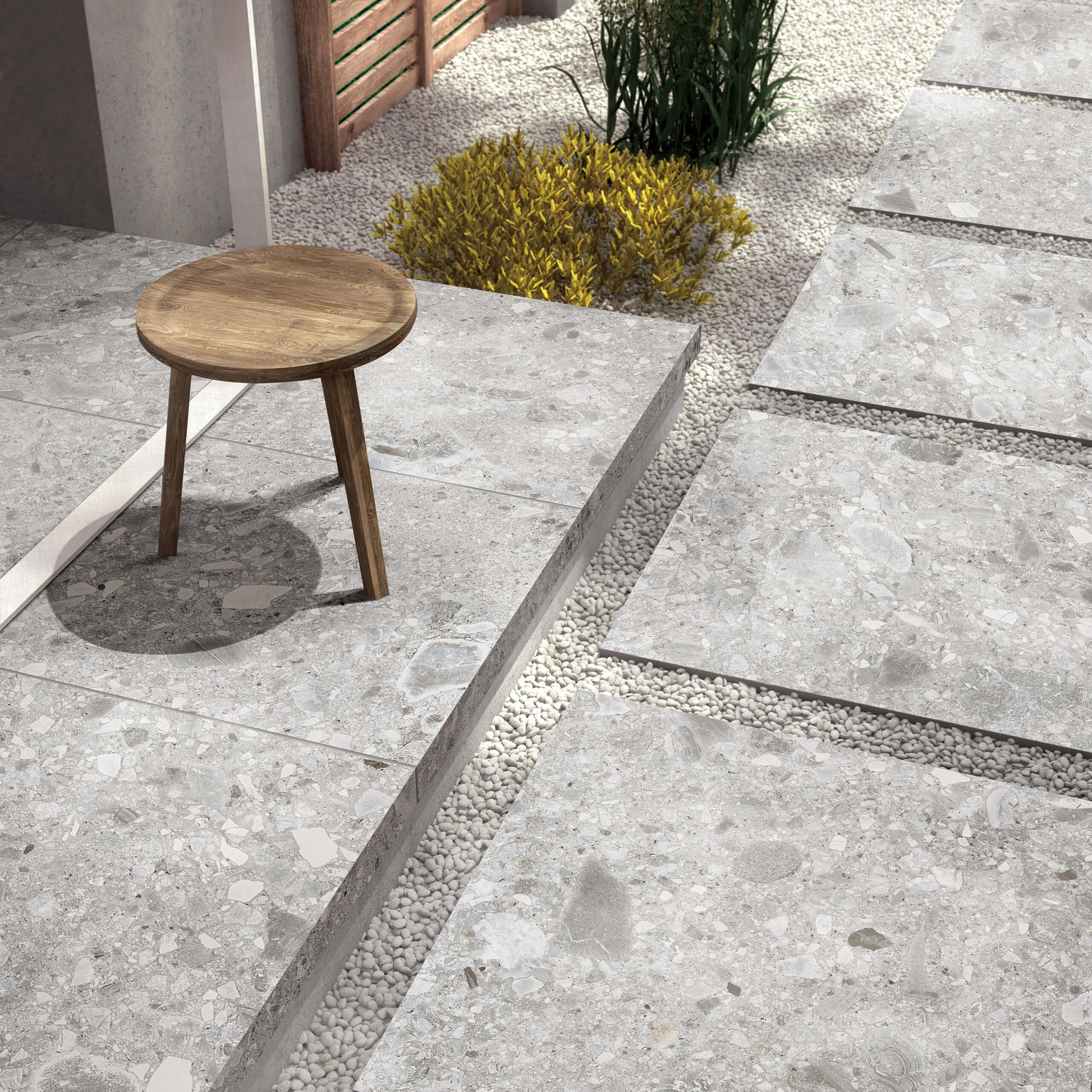 Ceppo Stone InOut 600x600mm grey terrazzo-effect porcelain tiles laid as raised outdoor patio stepping platforms separated by white pebble gravel channels, styled with a wooden stool and ornamental shrubs.