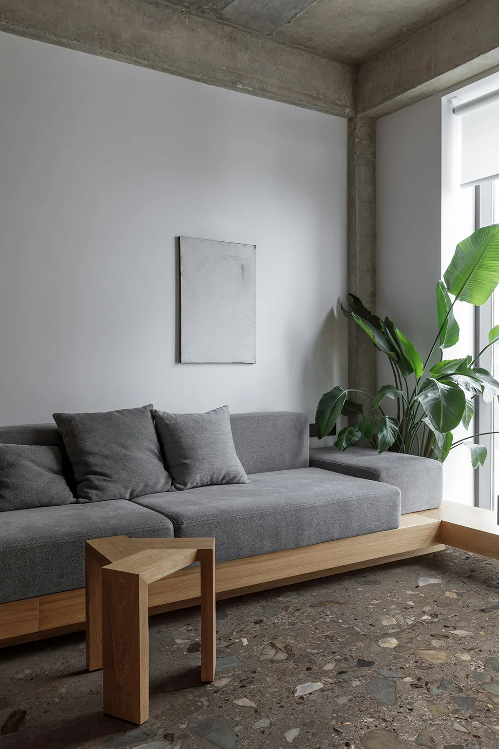 Ciottoli Sassi Antrac. 604x604mm terrazzo-style floor tiles in a minimalist living room with a grey sofa, timber frame, and indoor tropical plant.