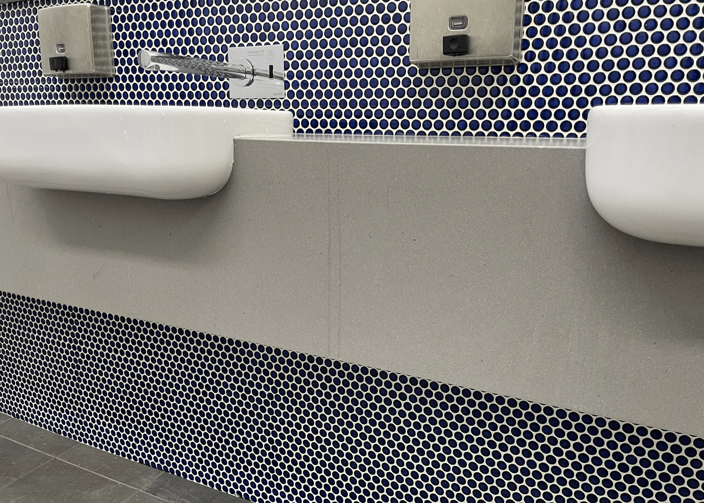 Cobalt Blue Gloss Diameter Glazed Porcelain Penny Round 19mm tiles installed on a public bathroom feature wall behind stepped grey stone vanity benchtops with wall-mounted faucets and soap dispensers.