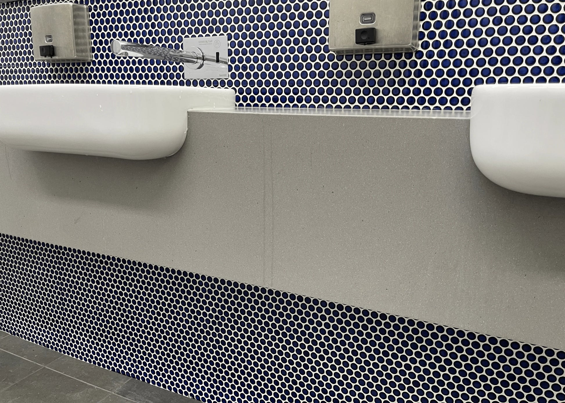 Cobalt Blue Gloss Diameter Glazed Porcelain Penny Round 19mm tiles installed on a public bathroom feature wall behind stepped grey stone vanity benchtops with wall-mounted faucets and soap dispensers.